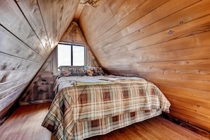 Ein gemütliches Schlafzimmer im Dachgeschoss mit Holzwänden und einer geneigten Decke, mit einer Karo-Bettdecke und einem Fenster mit Blick.