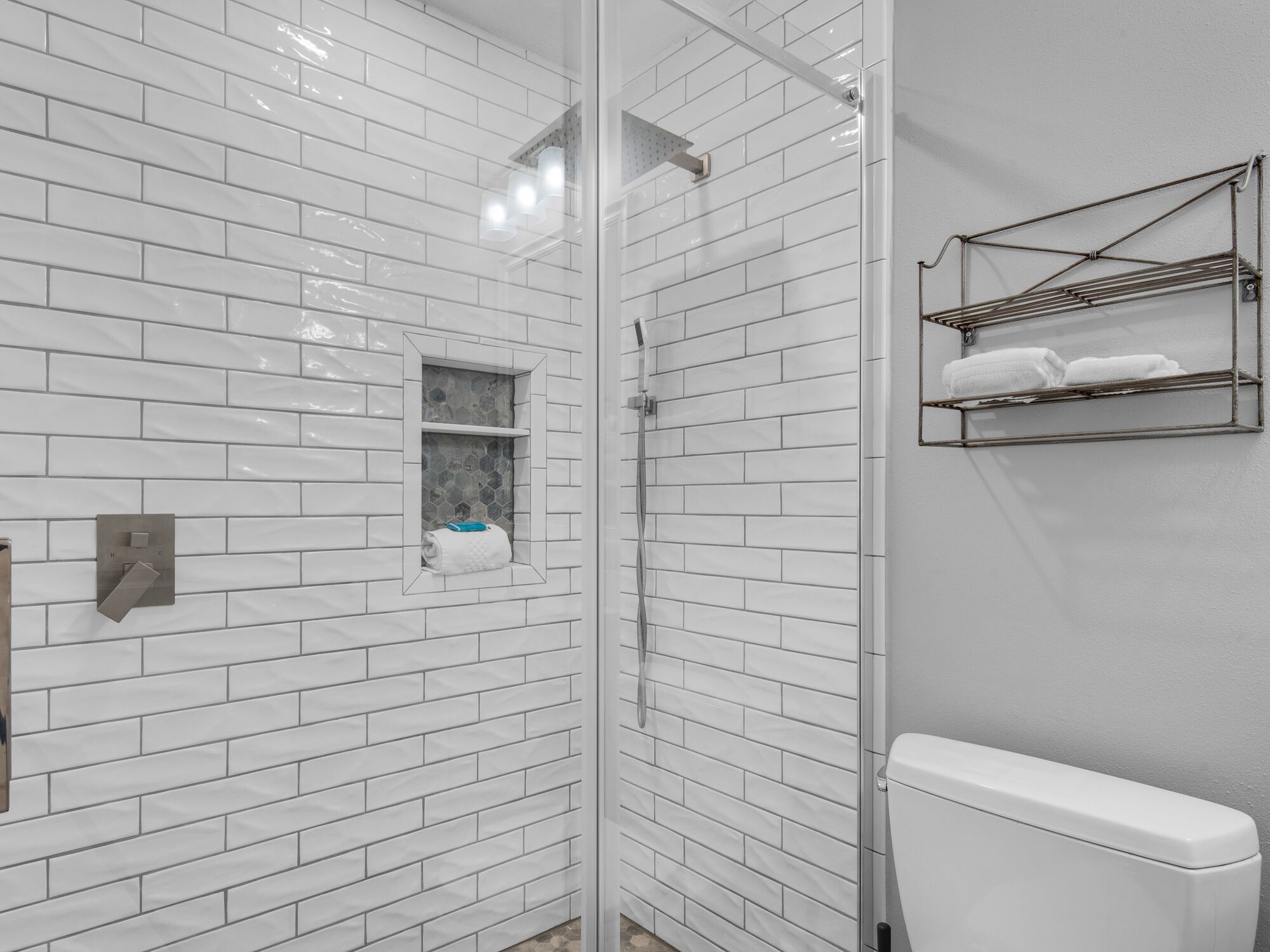 Ein modernes Badezimmer mit weißen, rechteckigen Fliesen, einer gläsernen Duschkabine, einer Toilette und einem Handtuchhalter mit gefalteten Handtüchern.