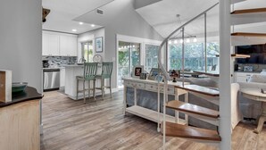 Een moderne keuken met witte kasten, een eethoek met houten stoelen, en een trap naar een woonruimte met een televisie en zitplaatsen.