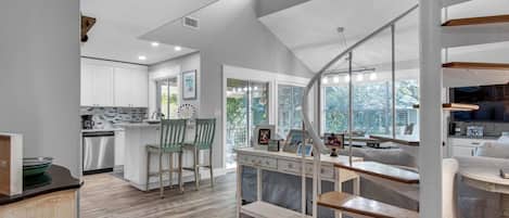 A modern kitchen with white cabinets, a dining area with wooden chairs, and a staircase leading to a living area with a TV and seating.