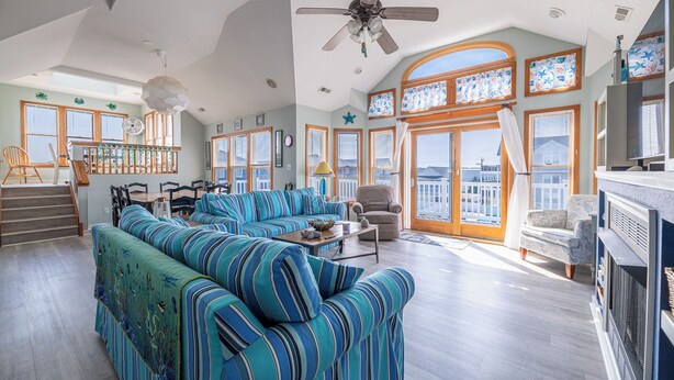 A spacious living room with a large sectional sofa, a ceiling fan, and a view of the outside through large windows. - The Life of Riley - NH MP 10.5, Ocean View, 265 Yds to Beach, Pool, Hot Tub, Pool Table, Flex Stays (Nags Head)