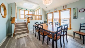 A dining area with a wooden table and chairs, a staircase with wooden railings, and large windows with a view. - The Life of Riley - NH MP 10.5, Ocean View, 265 Yds to Beach, Pool, Hot Tub, Pool Table, Flex Stays (Nags Head)