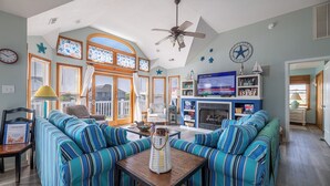 A living room with striped sofas, a ceiling fan, a fireplace, and a television. - The Life of Riley - NH MP 10.5, Ocean View, 265 Yds to Beach, Pool, Hot Tub, Pool Table, Flex Stays (Nags Head)