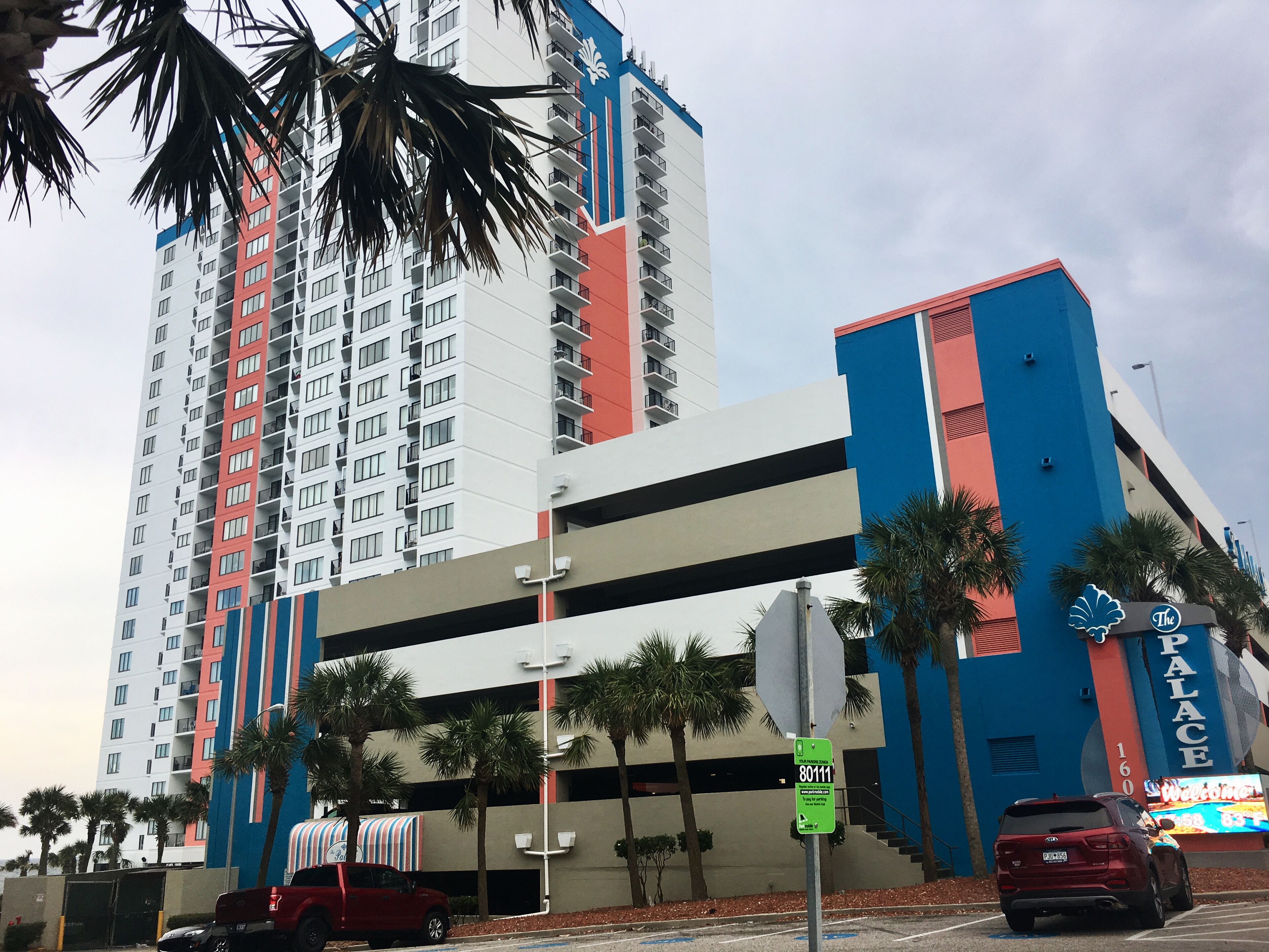 Un hotel de varios pisos con una fachada llamativa en rojo y azul, un aparcamiento y palmeras en primer plano.