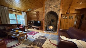 A cozy log cabin interior with a fireplace, wooden walls, a leather sofa, a television, and a bookshelf.