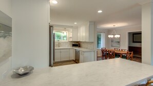 A modern kitchen with white cabinets, a dishwasher, and a dining area with a wooden table and chairs. - 20 Tupelo: Charming Sea Pines Home, Private Pool, Free Bikes (Hilton Head Island)