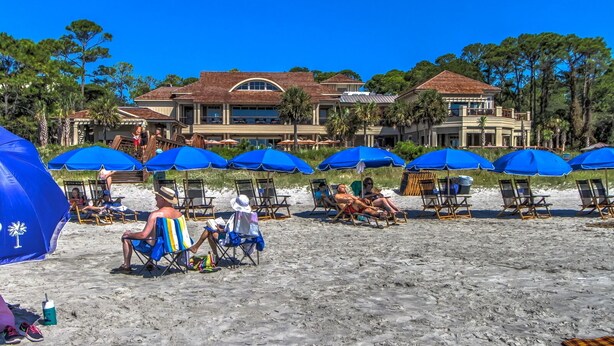 A beach with rows of blue umbrellas and beach chairs, a large building with a red roof in the background, and palm trees. - 20 Tupelo: Charming Sea Pines Home, Private Pool, Free Bikes (Hilton Head Island)