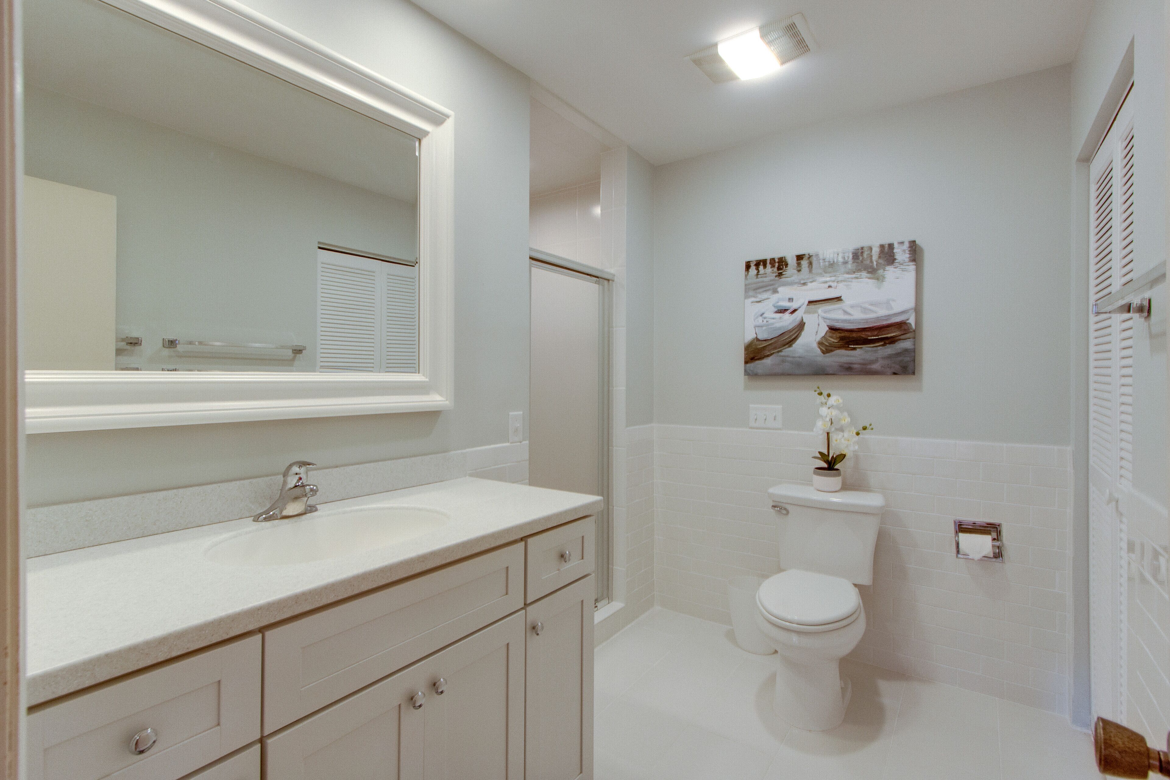 Une salle de bain moderne avec un grand miroir, des éléments d’ameublement blancs et un tableau encadré au mur.