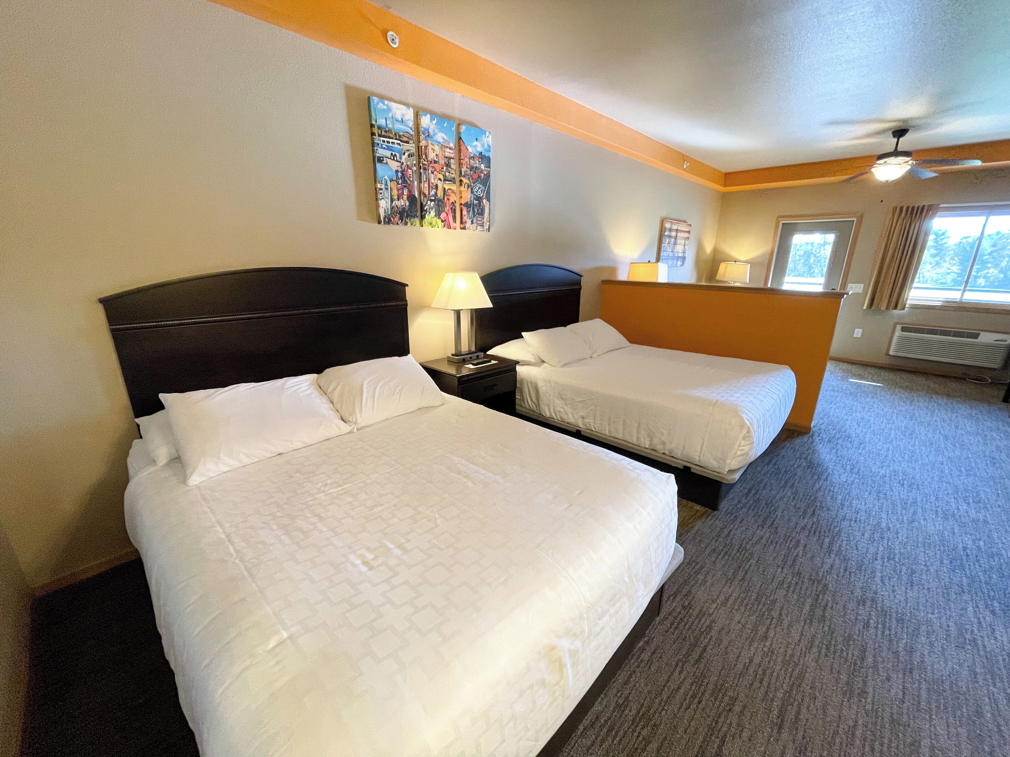 Een hotelkamer met twee bedden, een hoofdbord, een nachtkastje, een lamp, een plafondventilator, een raam met gordijnen en een schilderij aan de muur.