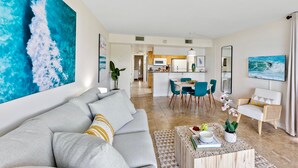 Uma sala de estar moderna com um sofá cinzento, uma mesa de centro com livros e uma planta, uma área de jantar com cadeiras azuis e uma cozinha ao fundo.