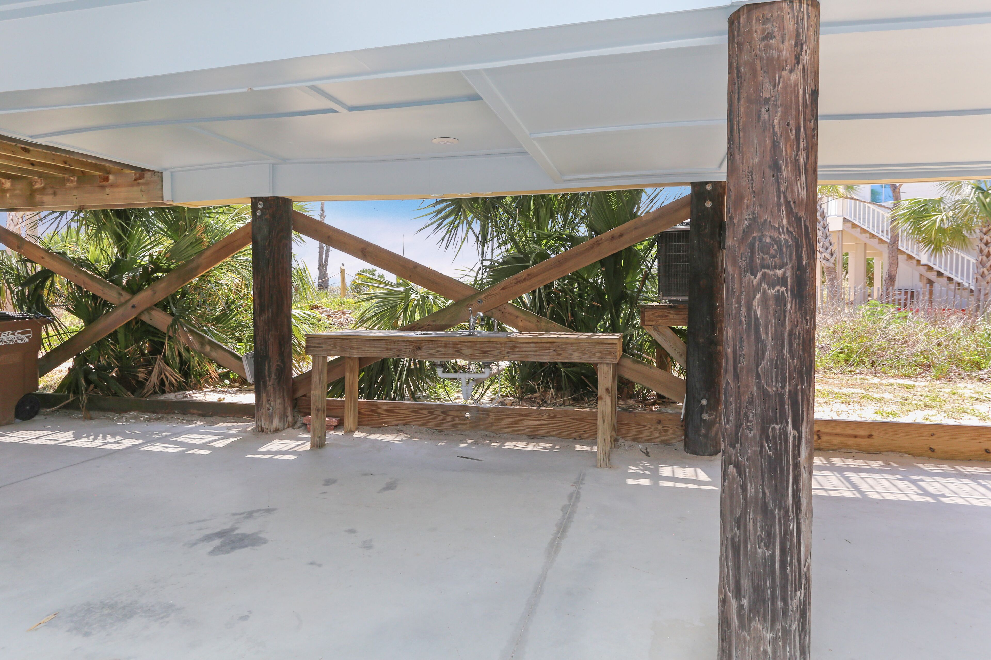 A construction site with wooden beams and a concrete floor, surrounded by palm trees and a building in the background.