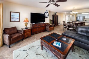 A living room with a flat-screen TV, a ceiling fan, a dining area with a kitchen in the background, and a large clock on the wall.