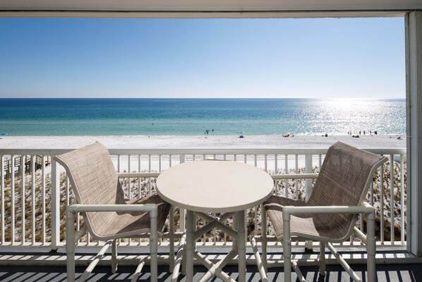 A balcony with two chairs and a table, overlooking a beach with people and a clear sky.