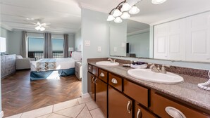 A modern bedroom with a large bed, a separate sitting area, and a bathroom with dual sinks and a large mirror.