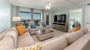 A spacious living room with a large sofa, two armchairs, a coffee table, and a television. There is a balcony with a view, and the room has a modern design with light blue walls and white ceilings.