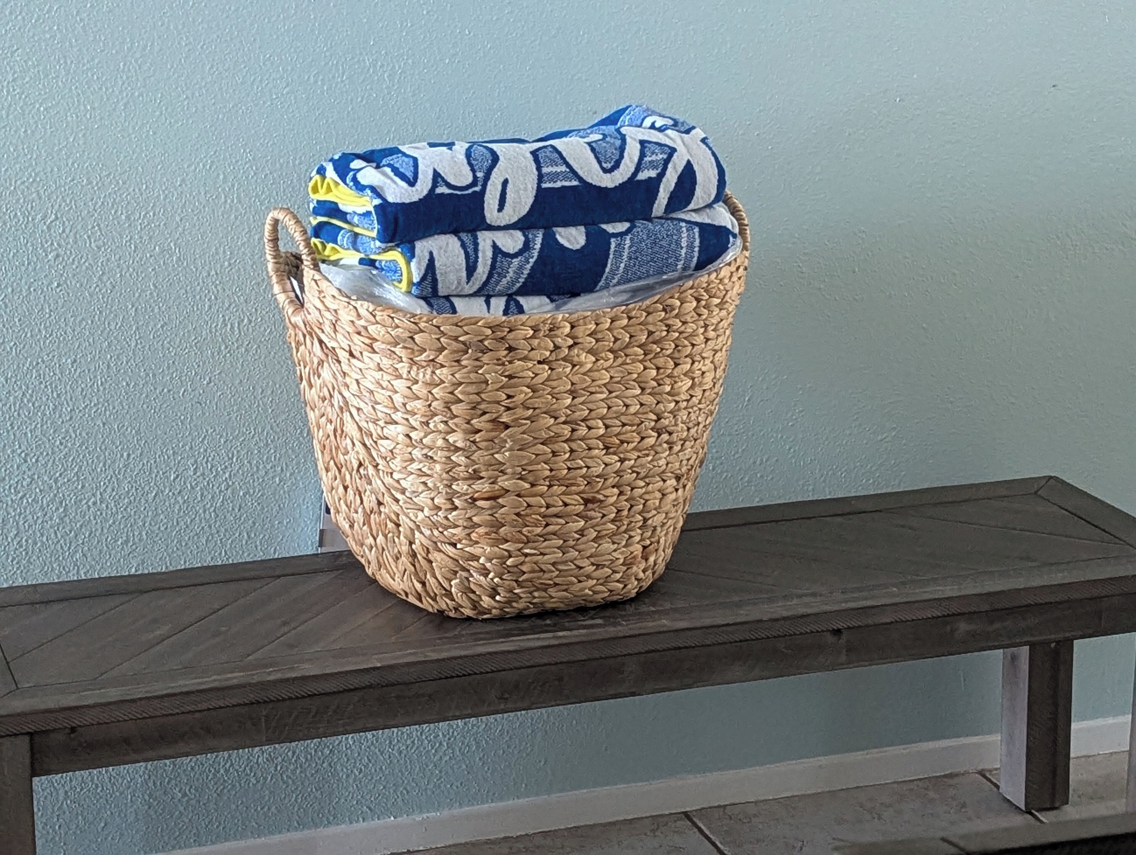 A woven basket with a blue and white patterned fabric inside, placed on a wooden bench against a light blue wall.