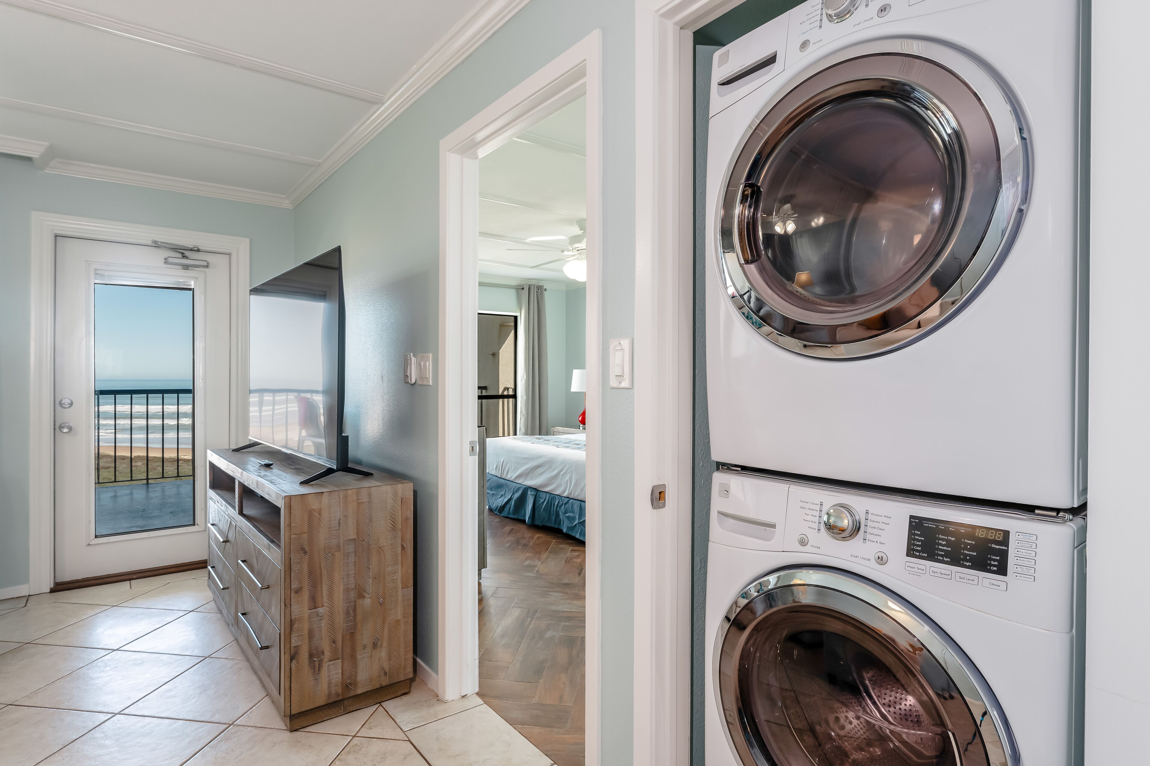 Une buanderie équipée d’une machine à laver et d’un sèche-linge superposés, d’un meuble en bois et d’une porte donnant sur une chambre avec un lit et une vue sur l’océan.
