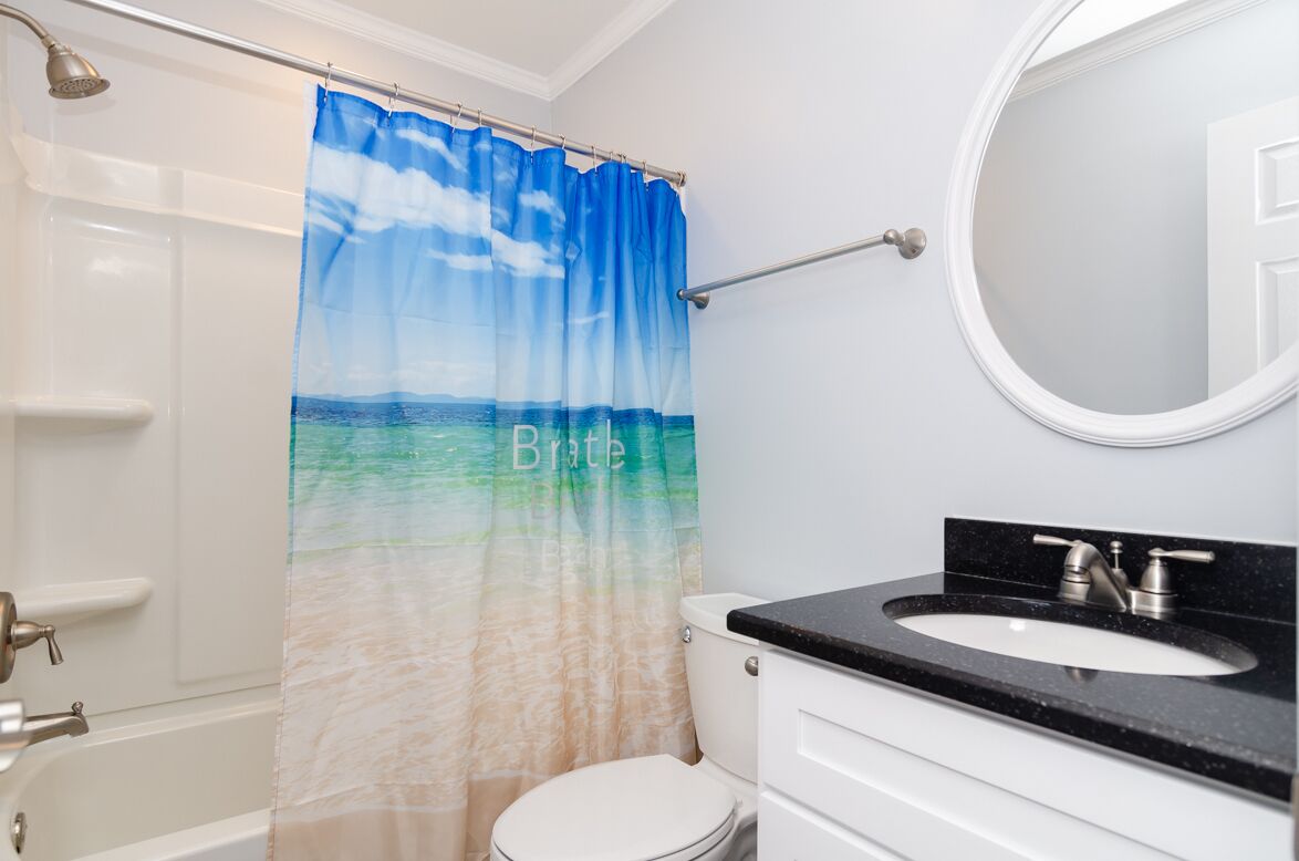 Ein Badezimmer mit Duschvorhang mit Strandmotiv, einer weißen Toilette, einem schwarzen Waschbecken mit Spiegel darüber und einer weißen Tür.