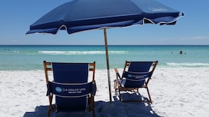 Two beach chairs with 'La Dolce Vita' branding and a blue umbrella on a sandy beach with the ocean in the background.