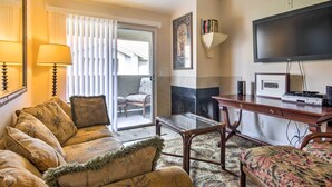 Un salon comprenant un canapé, un fauteuil et une table basse en verre. On y trouve également une cheminée, un téléviseur fixé au mur et une porte donnant sur un balcon.