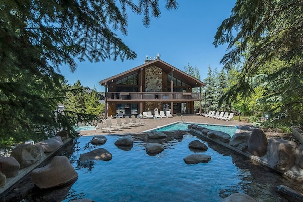 A lodge with a pool surrounded by rocks, a deck with lounge chairs, and a forested backdrop.