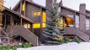 A modern multi-story house with a sloped roof, large windows, and a prominent evergreen tree in the front yard. - Canyons Resort Ski Home | Walk to Skiing + Free On Call Shuttle | 2 Fireplaces & 3 Cal-King Suites (Park City)