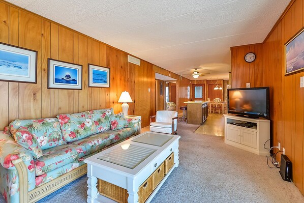A cozy living room with a floral sofa, a coffee table, and a television. There is a kitchen area with a dining table and chairs visible in the background. - Spacious, comfy 2 bedroom oceanfront condo with free WiFi, an outdoor pool, and a pretty ocean view located uptown just steps to the beach! (Ocean City)