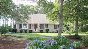 Ein zweistöckiges Haus mit einer Veranda, umgeben von einem gepflegten Rasen und einem Blumenbeet mit violetten Blüten.