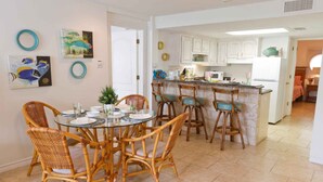 A dining area with a round glass table, wicker chairs, and a kitchen with white cabinets and a central island.