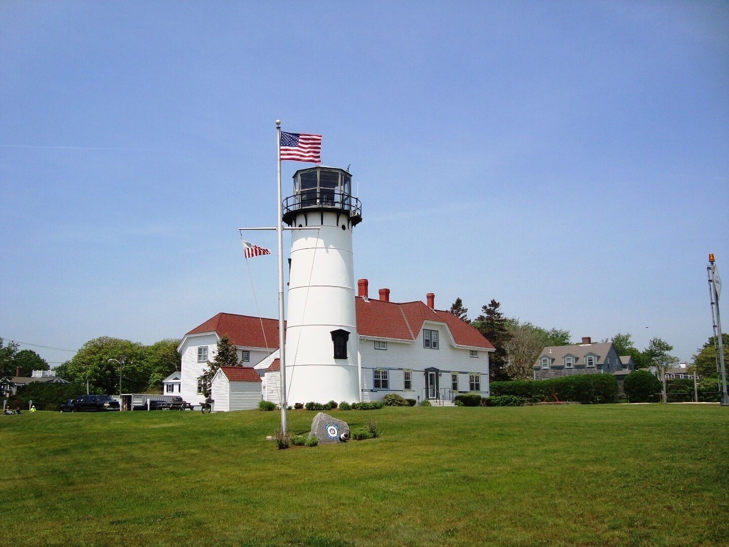A white lighthouse with a red roof, an American flag, and a nearby white building with a red roof.