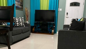 A living room with a flat-screen TV on a stand, two sofas, a dining table with chairs, and a door with a keypad lock. - Sleep Easy, Relax In Comfort (Caymanas Estates)