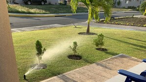 A patio area with a blue bench, a concrete pillar, and a lawn with small trees and a palm tree. - Sleep Easy, Relax In Comfort (Caymanas Estates)