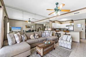 A living room with a sectional sofa, a coffee table, a television, and a kitchen area with cabinets and appliances.