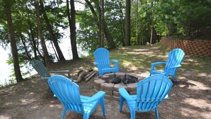 A fire pit with four blue Adirondack chairs arranged around it in a wooded area. - Spacious, waterfront cottage in Traverse City. Fireplace. Dog friendly. (Traverse City)