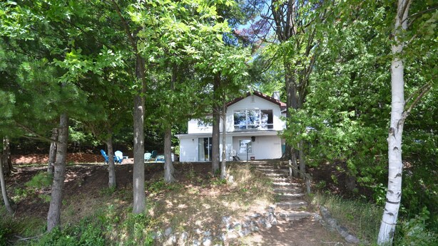 A two-story house surrounded by trees with a dirt path leading to the entrance. - Spacious, waterfront cottage in Traverse City. Fireplace. Dog friendly. (Traverse City)