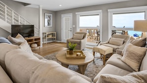Een moderne woonkamer met een grote beige bank, een salontafel en een televisie. Er is een balkon met buitenmeubilair en uitzicht op zee.