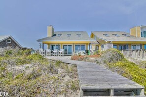 A beachfront property with a wooden boardwalk leading to a building with multiple windows and outdoor seating. - Dolphin Rise – Relaxation Starts Here (Topsail Beach)