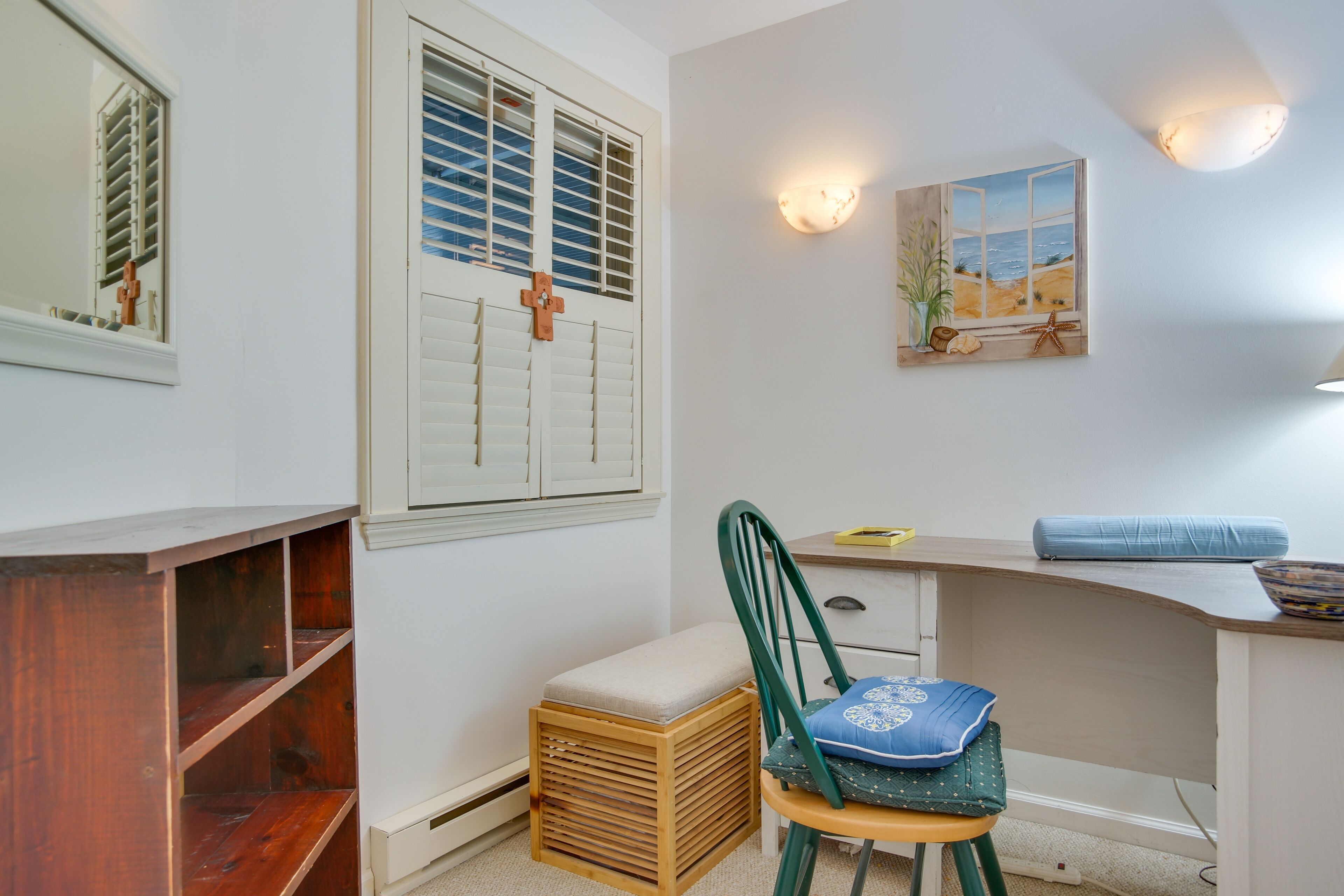 A room with a wooden desk, a chair, a small stool, a window with shutters, a wall-mounted shelf, and a painting of a beach scene.