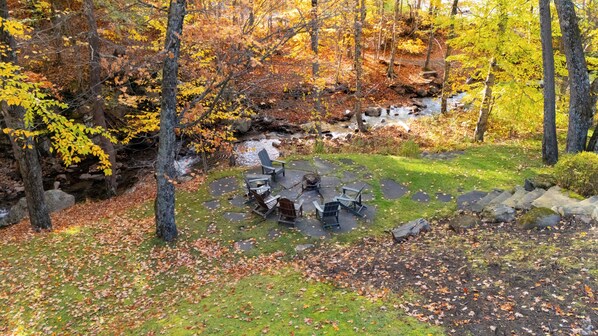 Un foyer circulaire en pierre, entouré de fauteuils Adirondack, se trouve dans une zone boisée près d’un ruisseau.
