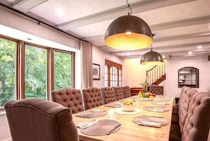 Une salle à manger avec une longue table, des chaises à revêtement marron, un luminaire suspendu et de grandes fenêtres donnant sur la forêt.