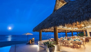 An outdoor dining area with thatched umbrellas, wooden tables, and chairs, overlooking a beach at night. - On 7th Hole Fairway with Access to Beach Clubs! (Punta Mita)