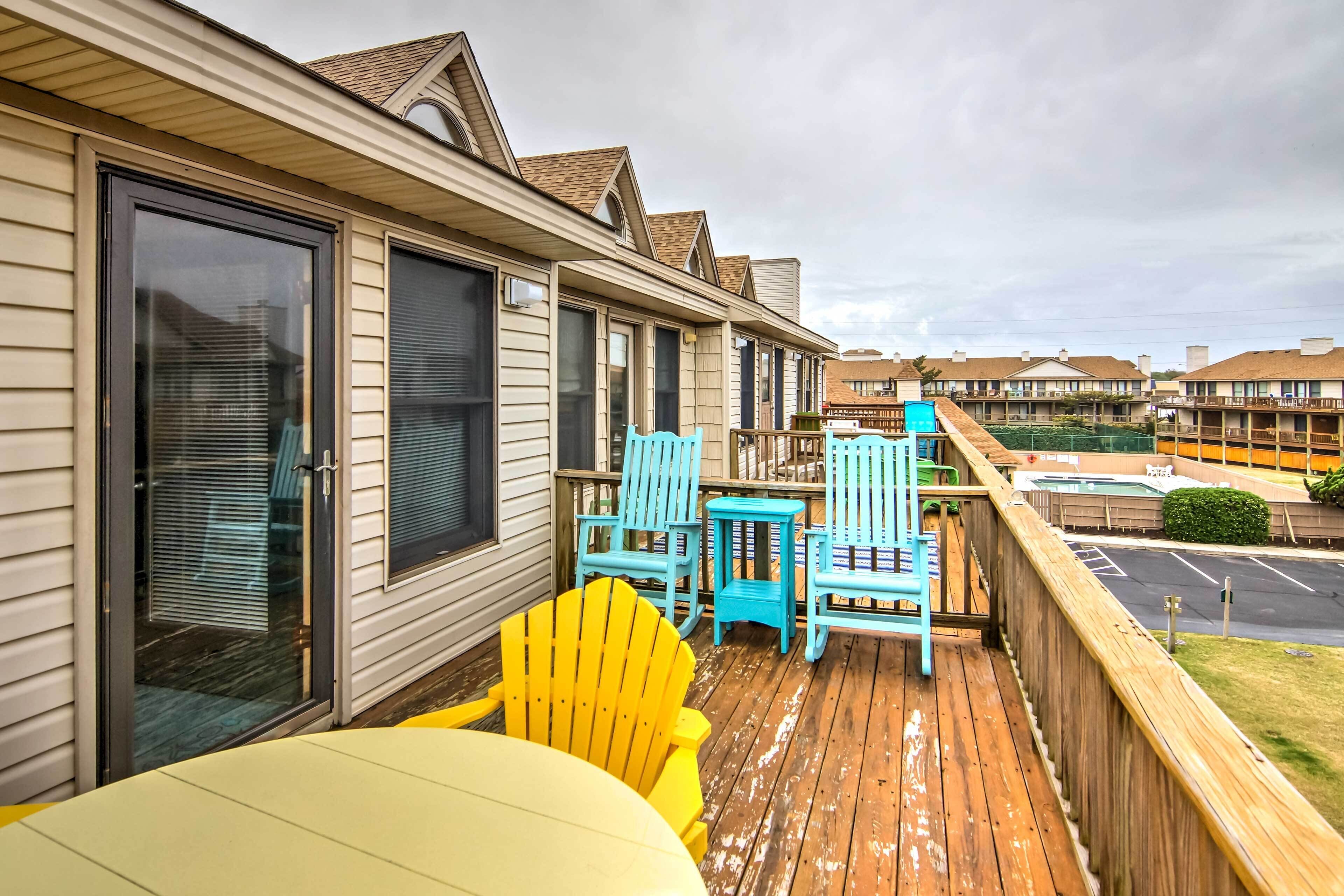 A wooden deck with yellow and blue Adirondack chairs, a small table, and a glass door leading to a covered area.
