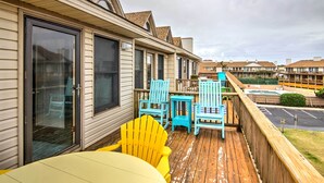 Een houten terras met gele en blauwe Adirondack-stoelen, een kleine tafel en een glazen deur naar een overdekt gebied.