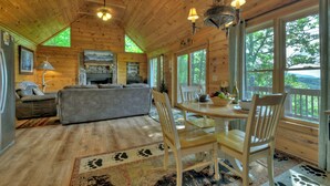 A cozy cabin interior with a wooden ceiling, a fireplace, a dining table with chairs, and large windows offering a view of the outdoors.
