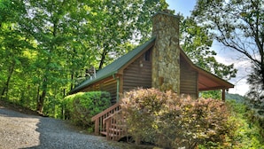 A log cabin with a stone chimney, surrounded by lush greenery and a gravel path leading to a wooden staircase.