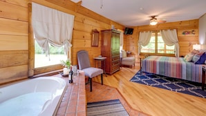 Interior d'una cabana de fusta amb un llit, una cadira, una taula amb una ampolla i vasos, un armari i un ventilador de sostre.