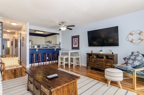 Un salon avec une table basse en bois, un téléviseur, un ventilateur de plafond et, au fond, une cuisine.