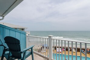 A balcony with a view of the ocean, a pool area with lounge chairs, and a blue building. - 1 Bedroom Oceanfront Condo in The Riggings! (Kure Beach)