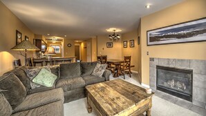 A cozy living room with a sofa, coffee table, fireplace, dining area, and kitchen in the background.