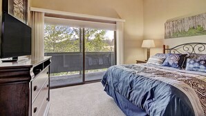 A bedroom with a large bed, a TV on a dresser, a balcony with a view of trees, and a painting on the wall.
